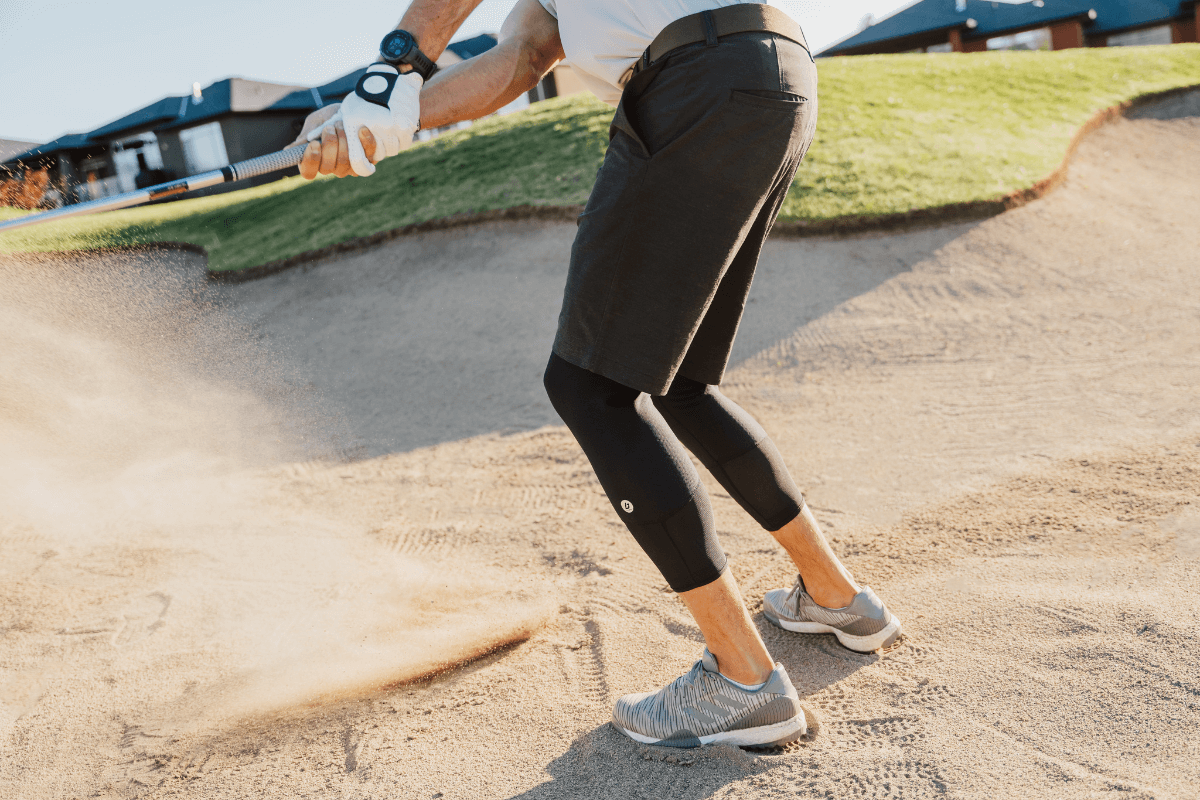 It's hard to dread chipping out of a sand trap when a golf game is supported by Bracelayer! In this photo, a man chips out of a sand trap with knees slightly bent in good golfing form, wearing Bracelayer's KS1 Vent knee stabilizing compression pants.