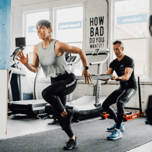 Two people in a gym, one person is running into a resistance band, the other is holding the resistance band behind her. Bracelayer Compression pants, compression tights, Exercises for knee strength, knee support, knees support, Vancouver knee brace, brace